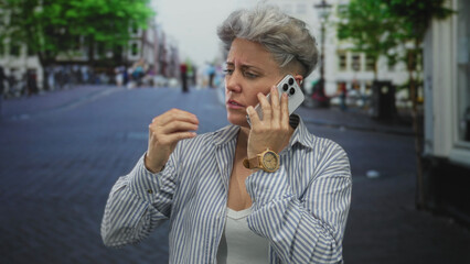Woman with short hair and furrowed brow holds smartphone in hand on crowded urban street; anger frustration.