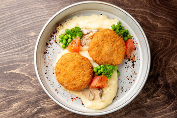 Breaded cutlets served over mashed potatoes with mushroom sauce, peas, tomatoes, and parsley in a white bowl on wooden background. Rustic comfort food presentation.