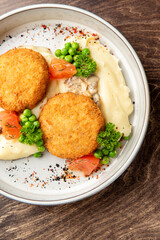Breaded cutlets served over mashed potatoes with mushroom sauce, peas, tomatoes, and parsley in a white bowl on wooden background. Rustic comfort food presentation.