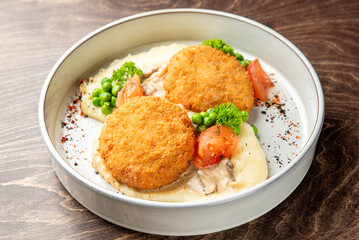 Breaded cutlets served over mashed potatoes with mushroom sauce, peas, tomatoes, and parsley in a white bowl on wooden background. Rustic comfort food presentation.