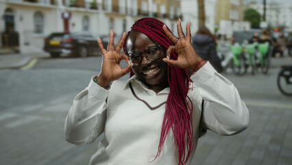 Woman on city street making ok gesture with fingers while smiling, wearing glasses and white top,...