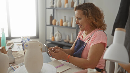 Artisan woman taps smartphone with fingers beside ceramic vase in studio; artistic joy quiet concentration.
