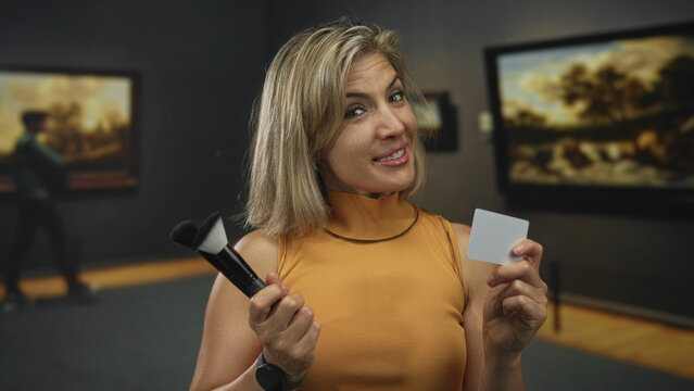 Woman holds makeup brush near face and grey card panel while smiling in art gallery; creativity curiosity. - Powered by Adobe
