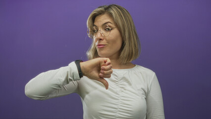 Young blonde woman wearing round glasses shows thumbdown gesture in purple studio setting;...