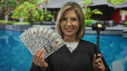 Young blonde woman judge stands smiling holding a fan of money and gavel by a pool at a building...