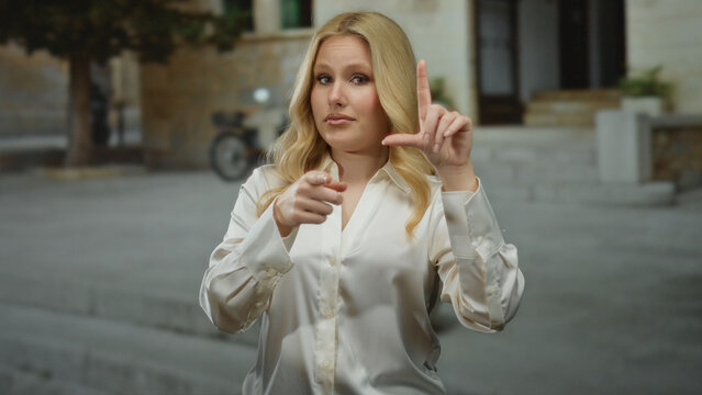 Young blonde woman making a looser gesture on a city street, wearing a white shirt with a neutral expression, pointing forward as if engaging in meaningful communication outdoors.