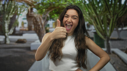 Obraz premium Young hispanic woman shows bare thumb in thumbs up gesture in green park outdoors under daylight; approval.