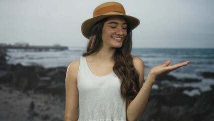 Young smiling hispanic woman wearing brown straw hat holds palm up on sunny beach; welcoming...