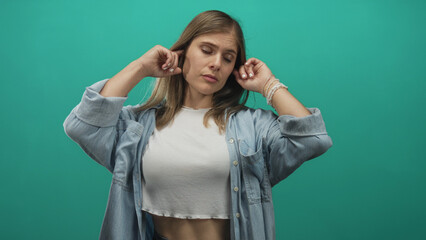 Blonde woman covers her ears with both hands while standing against a studio wall in turquoise...
