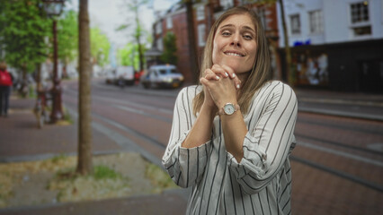 Woman smiles and clasps hands while standing on bustling city street lined with buildings and tram...