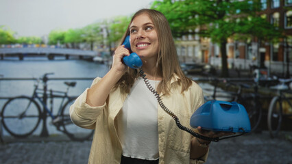 Young blonde woman holds blue telephone handset to ear on cobblestone street by canal bridge in...