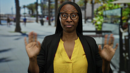 Woman with curly hair wearing glasses and yellow shirt expressing surprise outdoors on a sunny beach promenade with palm trees in the background, vibrant and lively atmosphere.