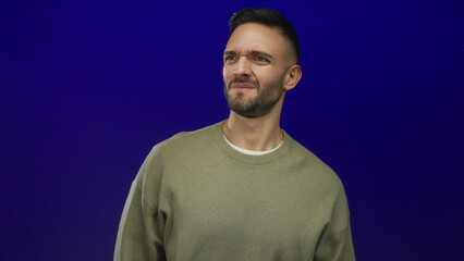 Hispanic man scrunching nose against blue background depicts displeasure due to bad smell, wearing...