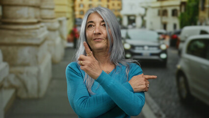 Fototapeta premium Woman with grey hair wearing blue sweater stands confidently on a bustling street, pointing with both hands, showcasing vibrant urban life and captivating expressions.