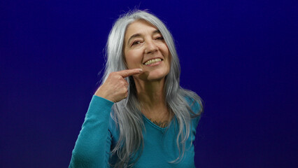 Fototapeta premium Senior woman with grey hair smiles warmly against an isolated blue background, pointing at her joyful expression with relaxed gestures, capturing a confident, serene moment.