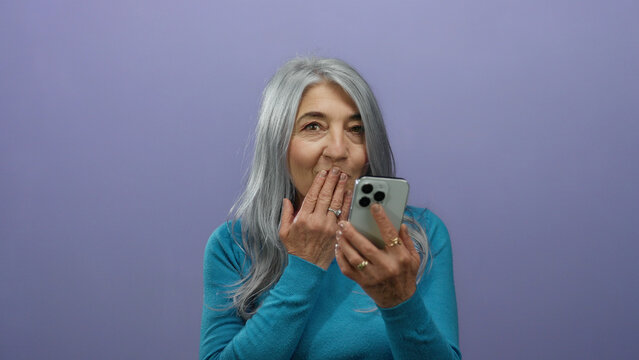 Senior woman with grey hair using smartphone against purple background, expressing surprise and amusement in isolated vibrant setting.
