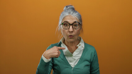 Woman with grey hair and glasses pointing at herself, wearing a green cardigan against an orange...