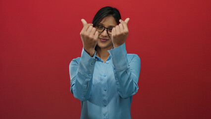 Woman smiling with glasses making a heart gesture with fingers against an isolated red background wearing a blue shirt indicating connection and warmth.