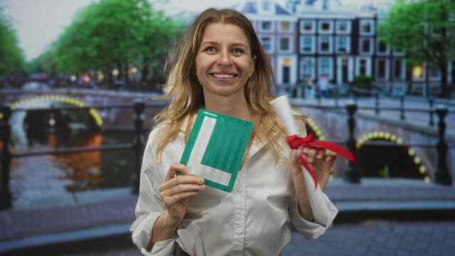 Woman smiles holding learner sign and rolled certificate with red ribbon on street by canal in amsterdam; pride celebration.