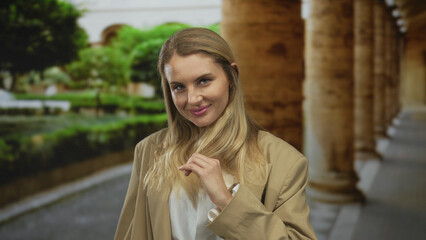 Woman smiling outdoors in a lush garden setting with arched architectural elements around, wearing...