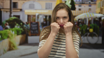 Fototapeta premium Woman in striped shirt raises fists in defensive boxing pose with serious expression on a busy street; defiance.
