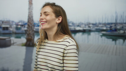 Woman smiling and showing teeth in white and brown striped top on street by a marina with boats and reflecting water; peaceful escape under soft daylight.