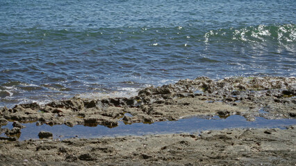 Fototapeta premium Mediterranean sea waves gently lap against rocky shore on a sunny day, highlighting the serene outdoor beach setting.