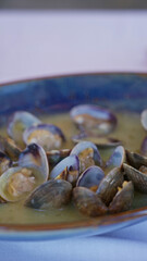 Hand holding spoon over bowl of clams in mediterranean sauce on restaurant table indoors with warm lighting creating cozy dining ambiance.