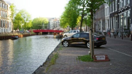 Blurred view of amsterdam canal with cars parked along the water and trees lining the walkway under a clear sky.