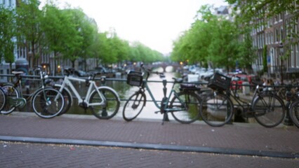 Bicycles lined along an amsterdam canal with a blurred bokeh effect highlighting classic dutch architecture and vibrant greenery on a serene city street.