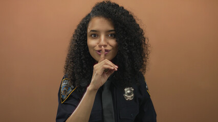 Young hispanic woman police officer in uniform gesturing silence with finger on lips against...