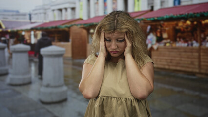 Woman with hands on temples at a busy street market near wooden souvenir stalls outdoors; stress shopping errands.