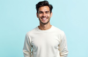 Obraz premium Young man with dark hair smiles broadly against a light blue studio backdrop. He wears a cream sweater, hands tucked in pockets, exuding confidence and good cheer.