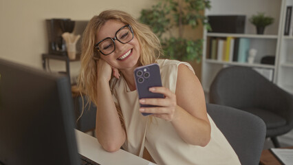 Woman wearing glasses holding smartphone, smiling while seated at a desk in an office interior beside a computer monitor; happiness work balance.