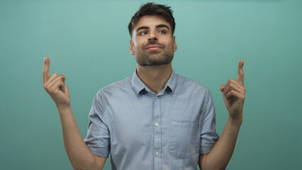 Man looking upward while pointing finger in studio against teal wall wearing light blue shirt;...
