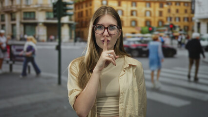 Fototapeta premium Woman with raised index finger pressed to lips gestures silence on a bustling city street, surrounded by blurred pedestrians; hush secrecy mystery calm.