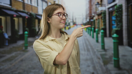 Fototapeta premium Blonde woman wearing glasses and beige shirt pointing finger on sunlit city street with cobblestone pavement and green bollards; confidence curiosity guidance direction.