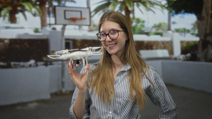 Fototapeta premium Young blonde woman wearing glasses holds drone near building wall outdoors; innovation exploration curiosity technology.