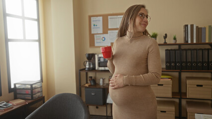 Pregnant woman enjoying coffee in an office setting, smiling and relaxed.