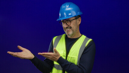 Man in hardhat and safety vest gesturing confidently against a blue background, depicting skilled professional engaging in a presentation or instruction setting.