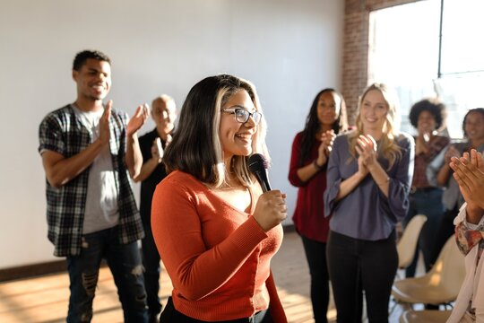 Group applauding a woman speaking with a microphone. Diverse audience clapping. Woman in focus, smiling, engaging with the crowd. Bright, positive atmosphere. Woman presenting in microphone at event.