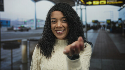Woman wearing cream sweater extends hand palm up and smiles in busy airport terminal; warm...