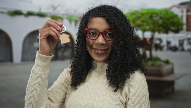 Black american woman wearing red glasses and a cream sweater holds a wooden house keychain with a bright smile on a bustling street; pride.