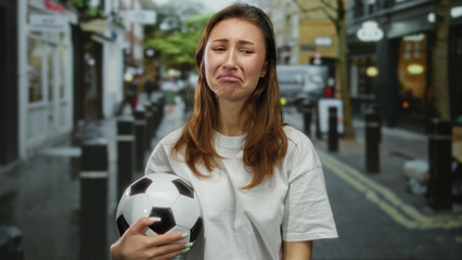 Woman in a city street holding a soccer ball with a sad expression, depicting emotion, outdoor scene, wearing casual white clothing, surrounded by urban cityscape.
