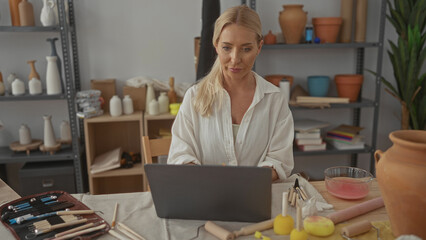 Woman types on laptop surrounded by pottery tools and clay jars under natural light in studio; creative concentration.
