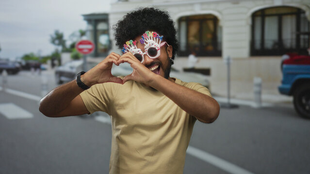 Man with birthday glasses smiling in a city street, making heart gesture, embodying joy and celebration in an urban outdoor setting. - Powered by Adobe