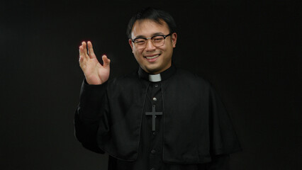 Asian priest man in glasses and black robe stands smiling against a dark background, holding hand up as if waving in a gesture of friendly greeting.