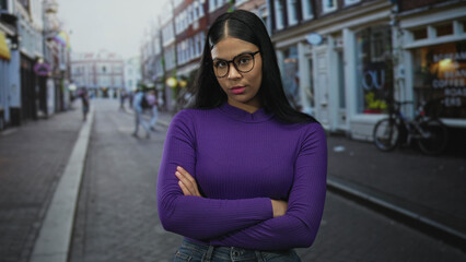Fototapeta premium Young latin woman wearing black eyeglasses with arms crossed on bustling urban street in daylight; calm reflection.