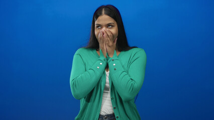 Woman wearing green cardigan covers mouth with both hands fully and closes eyes in blue studio;...