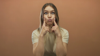 Blonde woman playfully puffing cheeks isolated against brown wall background wearing casual shirt...